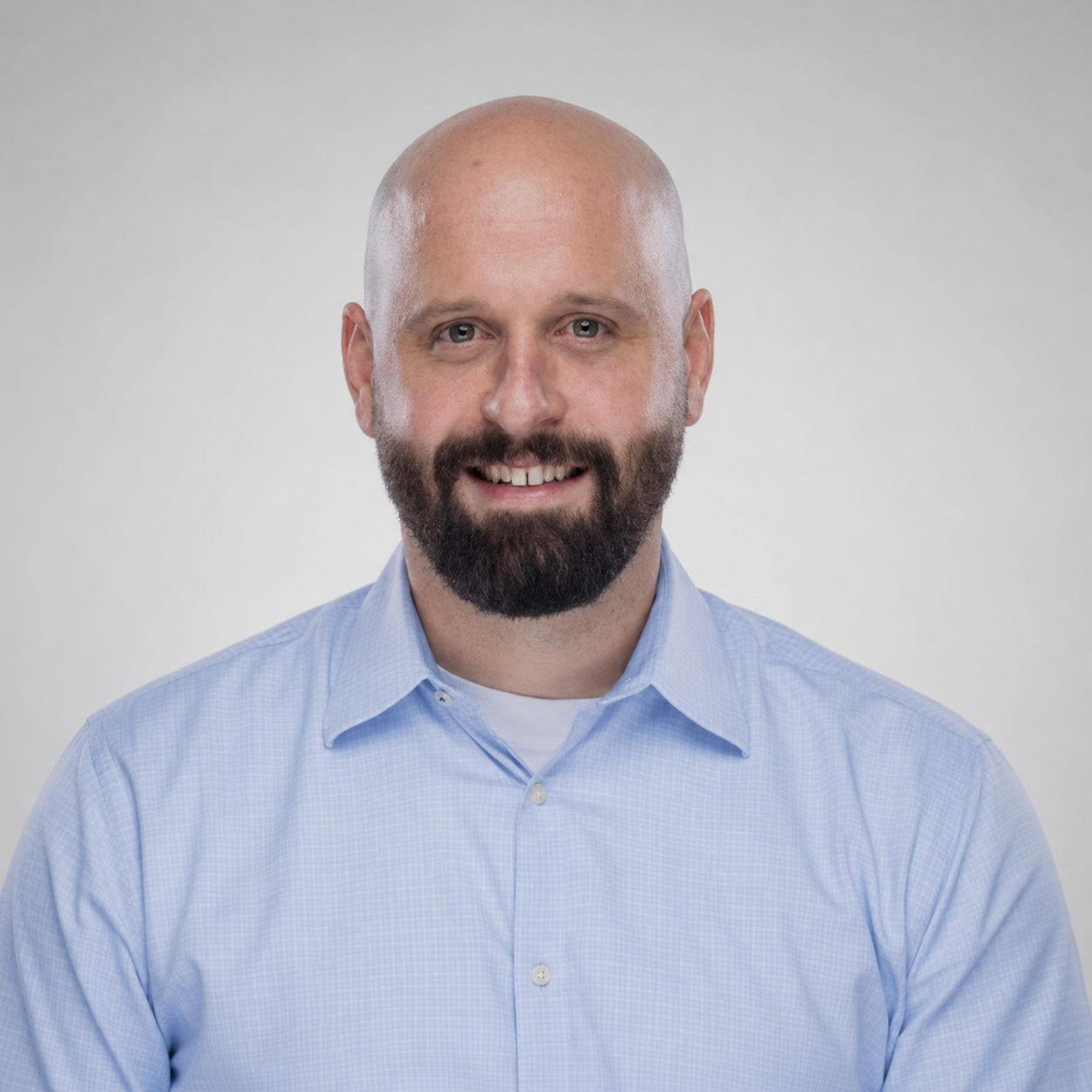 Carson Harnden - a bald man with a beard smiling at the camera wearing a blue collared shirt in front of a white background.